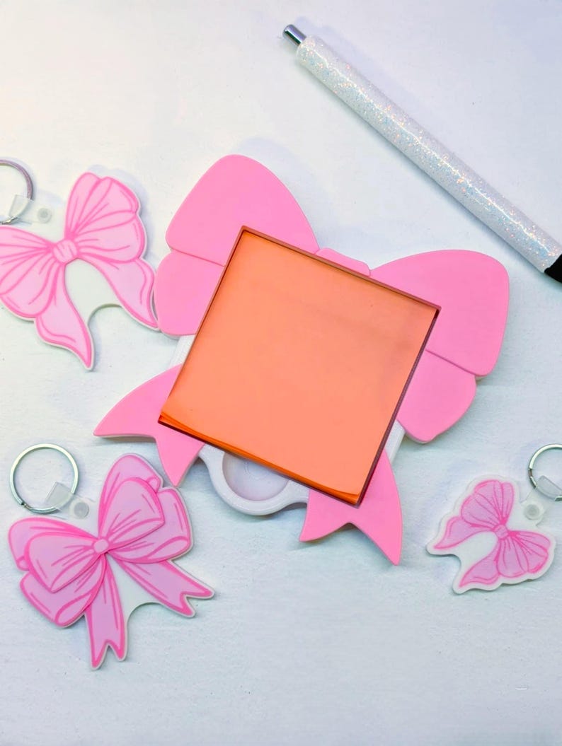 May include: A collection of pink bow-shaped keychains and a larger pink bow-shaped object with an orange square insert. A glittery pen rests nearby. The keychains and bow are on a white surface.