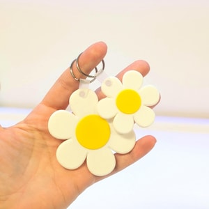Op de afbeelding: Twee sleutelhangers in de vorm van madeliefjes. Elke sleutelhanger heeft een witte bloem met een gele kern. De sleutelhangers worden bij elkaar gehouden door een zilveren metalen ring en een doorzichtige plastic band. De achtergrond is wit.