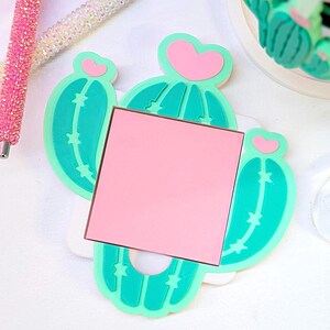 May include: A cactus-shaped memo holder with a pink square notepad. The holder is light green with pink heart accents. Two rhinestone pens are in the background, one pink and one white.