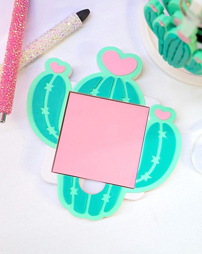 May include: A cactus-shaped memo holder with a pink square notepad. The holder is light green with pink heart accents. Two rhinestone pens are in the background, one pink and one white.
