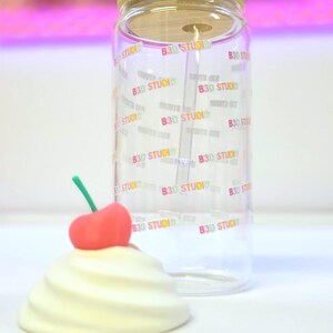 May include: A clear glass tumbler with a bamboo lid and a straw. The tumbler is printed with the text "B3D STUDIO" in a rainbow gradient. A decorative cream-colored swirl with a red cherry on top sits in front of the tumbler.