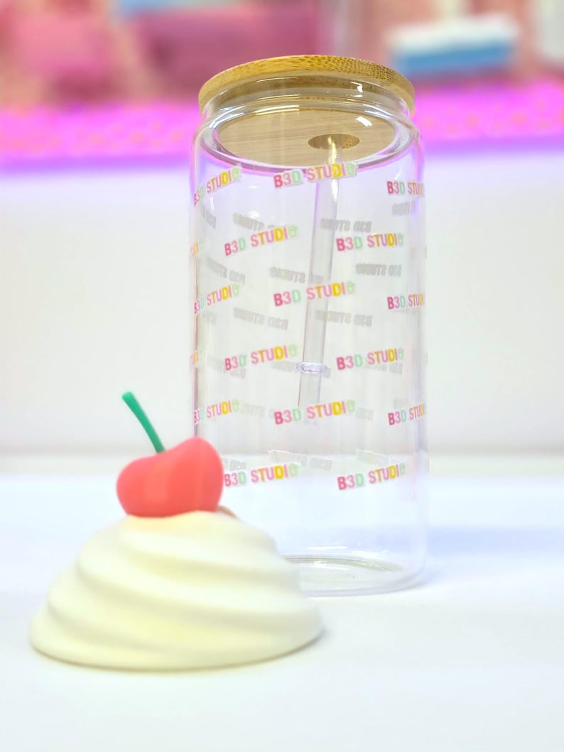 May include: A clear glass tumbler with a bamboo lid and a straw. The tumbler is printed with the text "B3D STUDIO" in a rainbow gradient. A decorative cream-colored swirl with a red cherry on top sits in front of the tumbler.