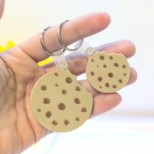 May include: Two cookie-shaped keychains. The keychains are tan with brown chocolate chips and a bite taken out of one. Each keychain has a silver ring and a clear plastic attachment.