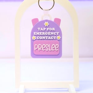 May include: A purple backpack-shaped keychain with the name "Preslee" on a pink rectangle. The keychain has the text "TAP FOR EMERGENCY CONTACT" and is decorated with yellow flower accents. It hangs from a metal ring.