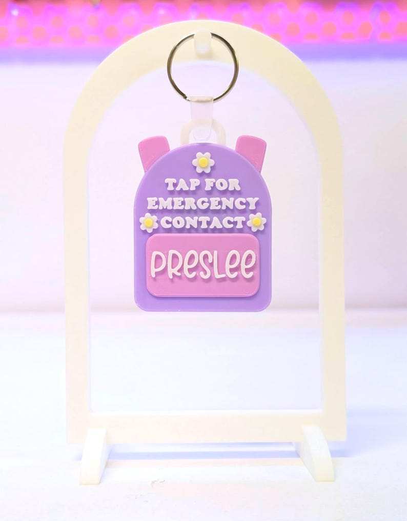 May include: A purple backpack-shaped keychain with the name "Preslee" on a pink rectangle. The keychain has the text "TAP FOR EMERGENCY CONTACT" and is decorated with yellow flower accents. It hangs from a metal ring.