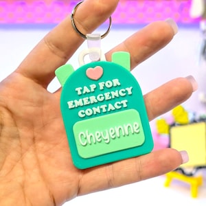 May include: A teal backpack-shaped keychain with the text "TAP FOR EMERGENCY CONTACT" and the name "Cheyenne" in white. A pink heart is above the text. The keychain is held by a silver ring.
