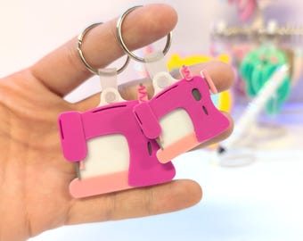 Peut inclure: Deux porte-clés en forme de machine à coudre rose avec des anneaux argentés. Les porte-clés ont une base blanche et un accent rose clair en bas. Les porte-clés sont tenus dans une main.