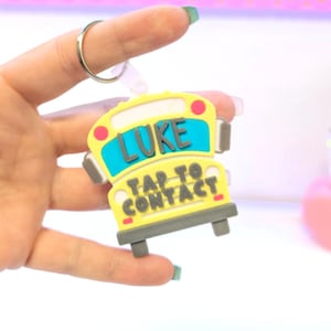 Könnte beinhalten: Ein gelber Schlüsselanhänger in Form eines Schulbusses mit dem Namen "LUKE" in Blau und dem Text "TAP TO CONTACT". Der Schlüsselanhänger hat rote und graue Akzente und einen silbernen Ring.