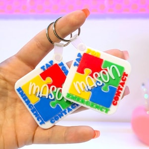 May include: Two white keychains with a puzzle piece design. Each keychain has the name "Mason" in white, surrounded by red, yellow, blue, and green puzzle pieces. The text "Tap for Emergency Contact" is also included.