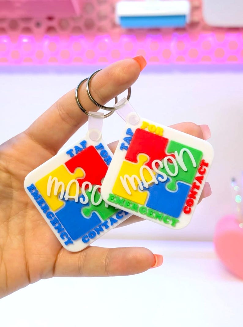 May include: Two white keychains with a puzzle piece design. Each keychain has the name "Mason" in white, surrounded by red, yellow, blue, and green puzzle pieces. The text "Tap for Emergency Contact" is also included.
