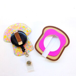 May include: Two novelty badge reels. One is a colorful, glittery shape with a black reel and a clear plastic strap. The other is a pink and brown bread-shaped design with a white strap. These are fun accessories for ID badges.