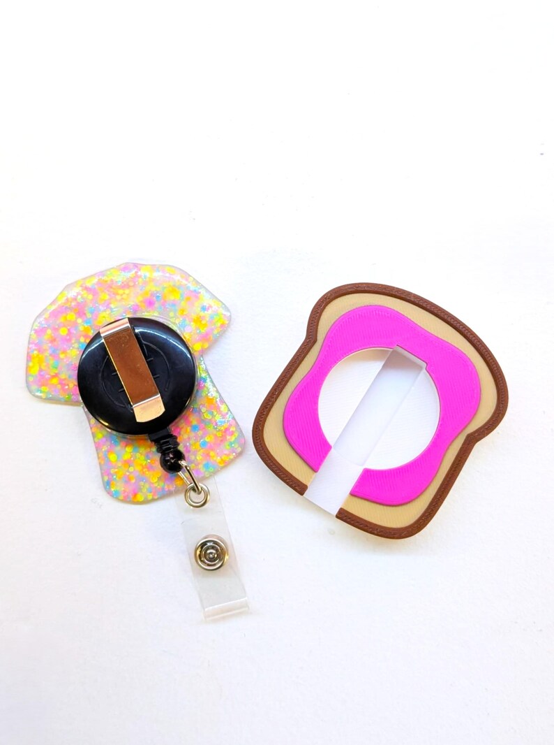 May include: Two novelty badge reels. One is a colorful, glittery shape with a black reel and a clear plastic strap. The other is a pink and brown bread-shaped design with a white strap. These are fun accessories for ID badges.