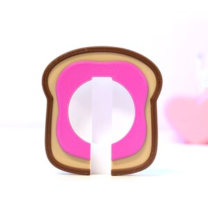 May include: A novelty bottle opener shaped like a slice of toast with a pink center. The opener has a brown crust, a tan middle layer, and a pink inner layer. The design is a playful take on a breakfast staple.