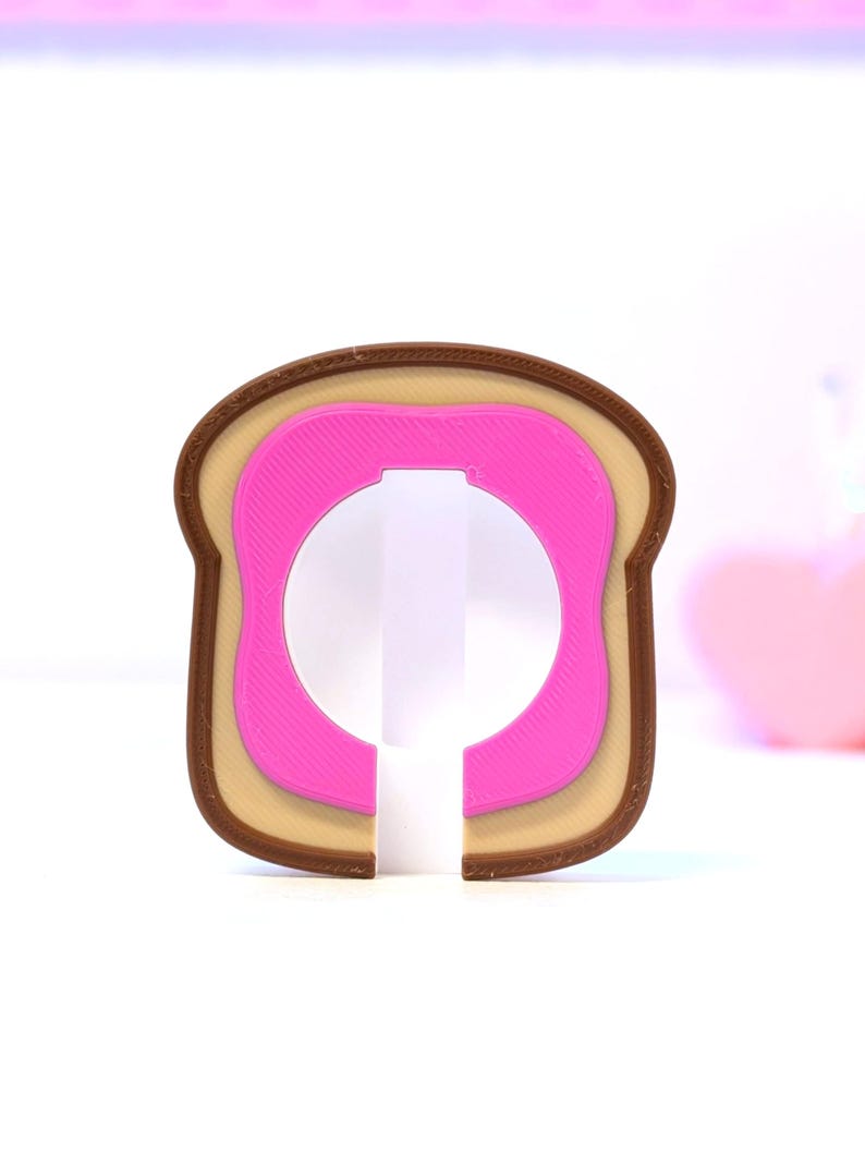 May include: A novelty bottle opener shaped like a slice of toast with a pink center. The opener has a brown crust, a tan middle layer, and a pink inner layer. The design is a playful take on a breakfast staple.