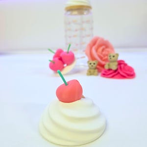May include: A whimsical arrangement featuring a white swirl-shaped object topped with a red cherry and green stem. Other elements include a clear bottle with a cream-colored lid, pink cherries, a pink rose, and tiny teddy bears.