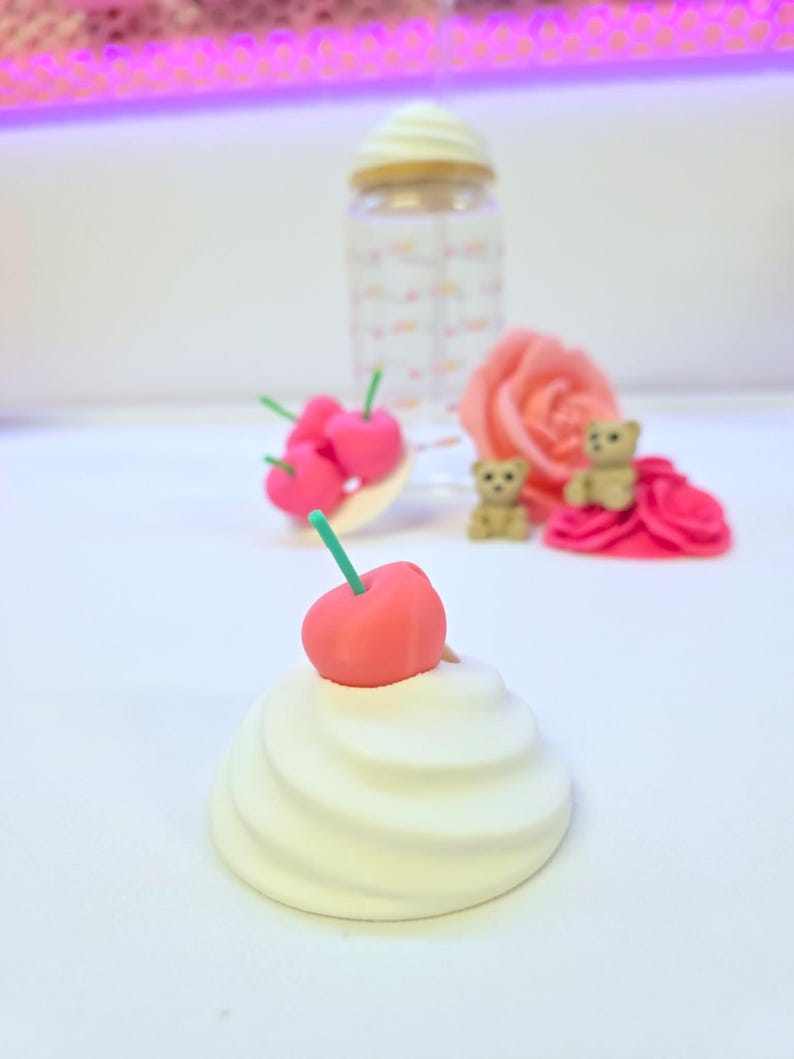 May include: A whimsical arrangement featuring a white swirl-shaped object topped with a red cherry and green stem. Other elements include a clear bottle with a cream-colored lid, pink cherries, a pink rose, and tiny teddy bears.