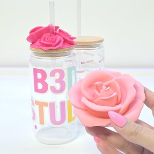 May include: Clear glass tumbler with a pink rose lid and a clear straw. The tumbler has the text "B30 STUDIO" in colorful letters. A pink rose is held in the hand, matching the lid's rose design.