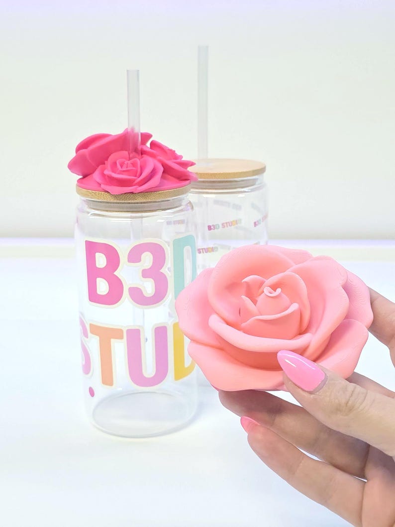 May include: Clear glass tumbler with a pink rose lid and a clear straw. The tumbler has the text "B30 STUDIO" in colorful letters. A pink rose is held in the hand, matching the lid's rose design.