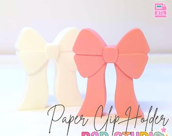 May include: Three 3D-printed bow-shaped paper clip holders in cream and coral colors. The image includes the text "Paper Clip Holder" and "B3D STUDIO". The top left corner reads "3D PRINT FILE" and "INSTANT DIGITAL DOWNLOAD".