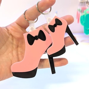 May include: Two pink high-heeled boot keychains with black bows and heels. The keychains are held up by silver key rings. The boots have a platform sole and a bow detail. The background is blurred.