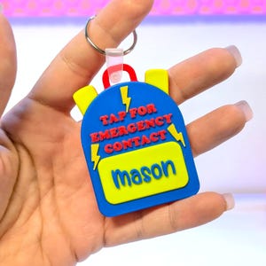 May include: A blue backpack-shaped keychain with yellow accents and the name "Mason" on a yellow rectangle. The keychain has the text "TAP FOR EMERGENCY CONTACT" and lightning bolt graphics in yellow and red.