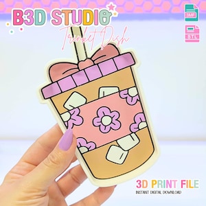 Peut inclure: Un vide-poches en forme de boisson glacée de dessin animé, avec un nœud rose, un motif floral rose et blanc, et le texte "B3D STUDIO Trinket Dish". Le plat est un fichier d'impression 3D.