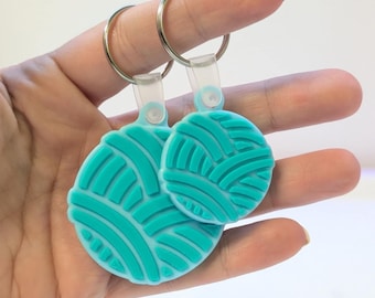 Peut inclure: Deux porte-clés turquoise, chacun avec un motif rond ressemblant à une pelote de laine. Les porte-clés ont des anneaux argentés et des attaches en plastique transparent. Le fond est blanc uni.