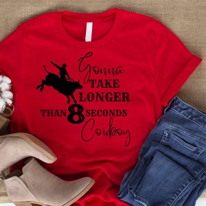 Puede incluir: Camiseta roja con una silueta negra de un vaquero montando un toro. El texto dice "Gonna take longer than 8 seconds cowboy".
