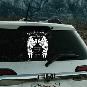 May include: A white car with a black rear window. The rear window has a white decal with a pair of angel wings and the text "In loving memory Brian Richards 3.15.67 Your wings were ready, but our hearts were not. 5.27.22". The car has a GMC logo and a California license plate.