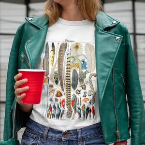 May include: A woman wearing a green beanie, a white t-shirt with a vintage illustration of feathers and birds, a green leather jacket, and denim shorts. The t-shirt has the word "Plumes" printed on it.
