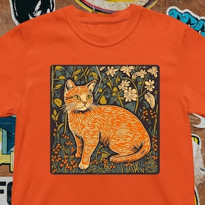 Op de afbeelding: Oranje t-shirt met een vierkante afbeelding van een oranje kat omringd door bloemen en bladeren. De kat zit, en het ontwerp heeft een vintage, illustratieve stijl. Het t-shirt heeft een heldere, gedurfde kleur.