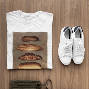 May include: White t-shirt with a bread graphic, olive green sweatpants, white sneakers, and maroon headphones on a wooden surface. The t-shirt features four different types of bread. The sneakers have white laces and navy blue accents.
