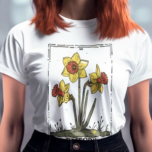 May include: A white t-shirt featuring a vintage botanical illustration of three yellow daffodils with orange centres. The illustration is framed in a black rectangle with the word "DAFFODIL" printed at the bottom.