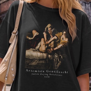 May include: A black short-sleeved t-shirt featuring a graphic print of the painting Judith Slaying Holofernes by Artemisia Gentileschi, with the text Artemisia Gentileschi Judith Slaying Holofernes 1620 printed in white below the artwork.