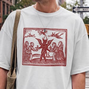 May include: A white short-sleeve t-shirt featuring a rectangular dark red graphic print of a central winged horned figure surrounded by four human figures and three flying creatures in a woodcut style, paired with a brown canvas shoulder bag.