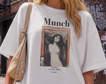 May include: A white short-sleeved t-shirt featuring a graphic print of Edvard Munch's Madonna painting. The text reads Munch, Madonna (ca. 1893-1894), and TASCHEN. The shirt has a loose fit and is paired with a tan shoulder bag and dark trousers.