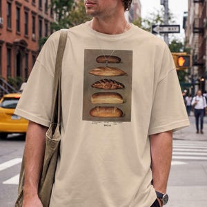 May include: A beige short-sleeve t-shirt featuring a rectangular graphic print of five different types of bread stacked vertically. Below the bread illustrations, the text reads BREAD in a simple, dark, capitalised font on the centre chest.