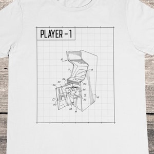 May include: White t-shirt with a black and white illustration of an arcade game with the text "PLAYER - 1" on a grid background.