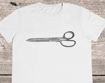 May include: A white short-sleeve t-shirt featuring a centered graphic illustration of a pair of metal scissors. The scissors are depicted in a horizontal orientation with two circular finger loops on the right side and a pointed blade on the left.