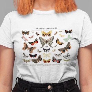 Puede incluir: Camiseta blanca con una ilustraci&oacute;n vintage de una colecci&oacute;n de mariposas. La ilustraci&oacute;n est&aacute; en tonos de marr&oacute;n, naranja, amarillo, blanco y negro. El texto "SCHMETTERLINGE II." est&aacute; impreso sobre la ilustraci&oacute;n.