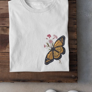 Puede incluir: Camiseta blanca con un gr&aacute;fico de mariposa amarilla y negra en el lado izquierdo. La mariposa est&aacute; rodeada de flores rosas y blancas.
