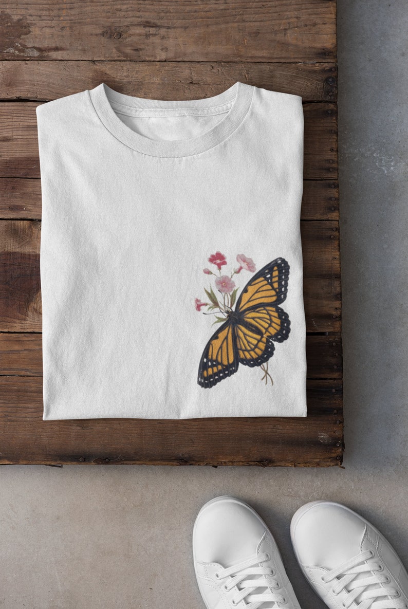 Puede incluir: Camiseta blanca con un gr&aacute;fico de mariposa amarilla y negra en el lado izquierdo. La mariposa est&aacute; rodeada de flores rosas y blancas.