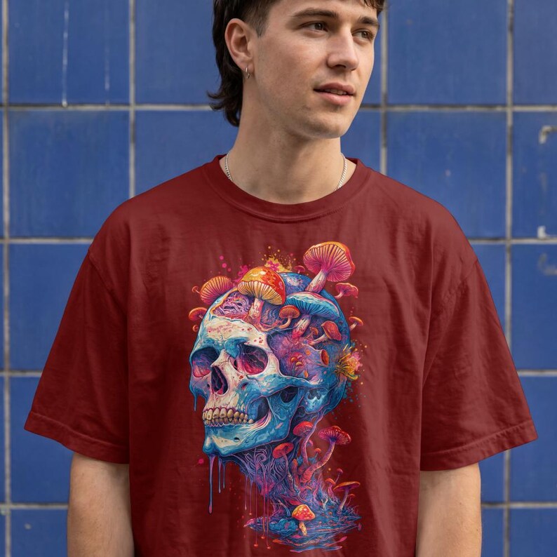 Op de afbeelding: Een bordeauxrood t-shirt met korte mouwen en een grafische print van een blauwe en paarse schedel met diverse kleurrijke paddenstoelen die uit de bovenkant en basis groeien. Het ontwerp bevat levendige rode, oranje en roze accenten.