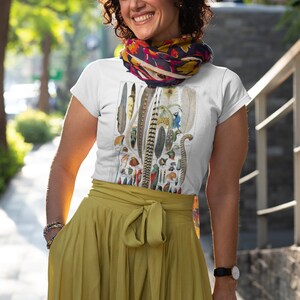 May include: A white t-shirt with a vintage illustration of a collection of feathers. The shirt is worn by a person with curly brown hair and a colorful scarf around their neck. The person is standing on a sidewalk.