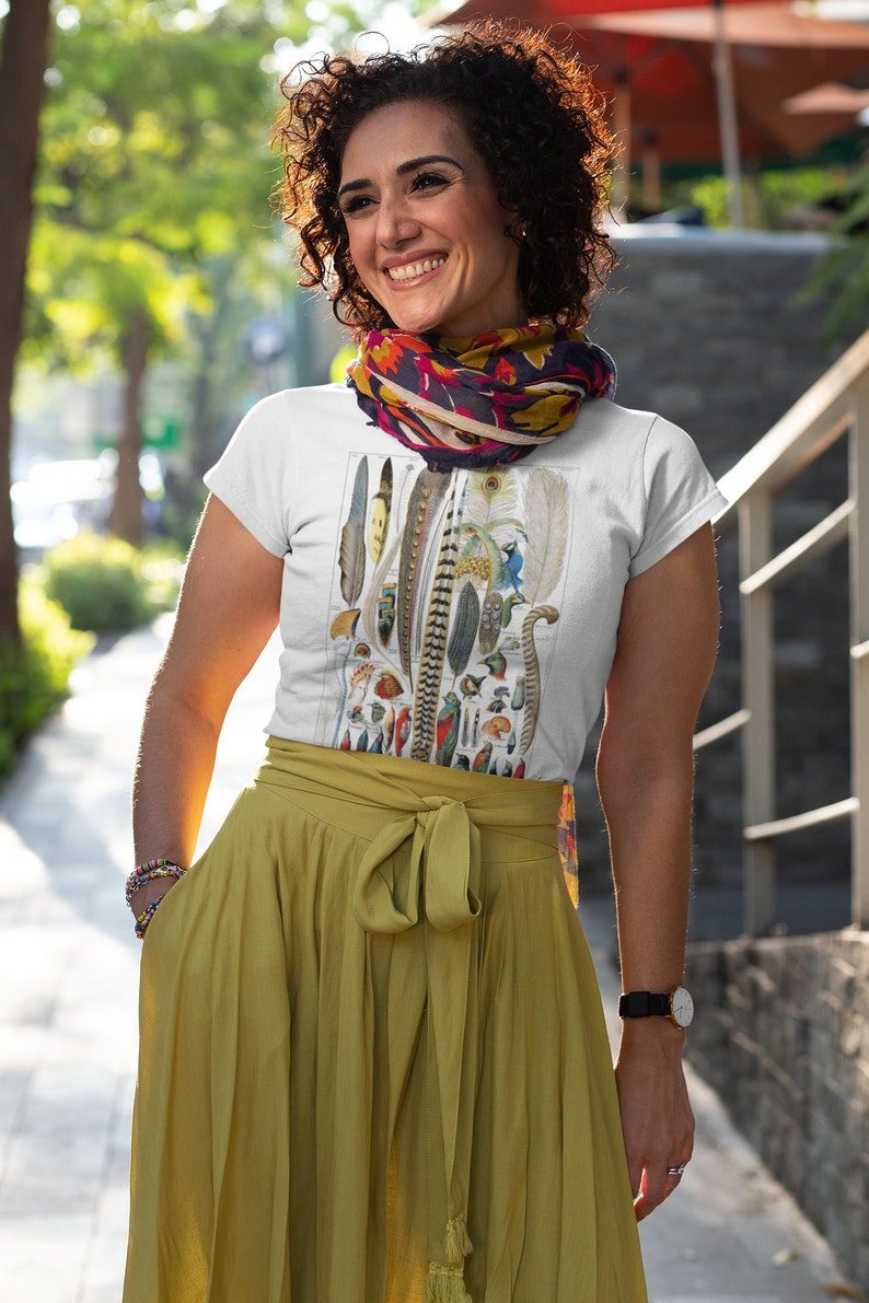 May include: A white t-shirt with a vintage illustration of a collection of feathers. The shirt is worn by a person with curly brown hair and a colorful scarf around their neck. The person is standing on a sidewalk.