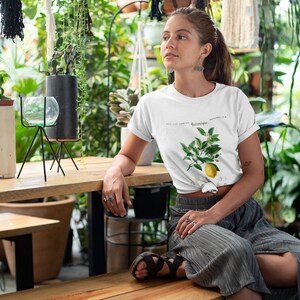 May include: A white short-sleeved t-shirt featuring a botanical illustration of a lemon branch with green leaves, white blossoms, and a single yellow lemon, accompanied by the text Botanique printed in a black serif font on the chest.