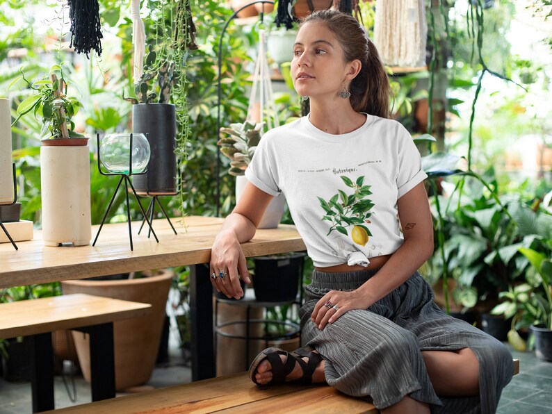 May include: A white short-sleeved t-shirt featuring a botanical illustration of a lemon branch with green leaves, white blossoms, and a single yellow lemon, accompanied by the text Botanique printed in a black serif font on the chest.