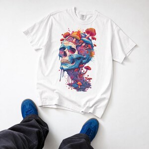 Op de afbeelding: Een wit t-shirt met korte mouwen en een kleurrijke grafische print van een menselijke schedel in levendige blauwe, paarse en roze tinten. De schedel is versierd met diverse oranje en rode paddenstoelen, met verfachtige druppels die naar beneden lopen.