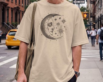 May include: A beige short-sleeve t-shirt featuring a black ink-style graphic of a crescent moon with a human face profile surrounded by various star shapes. The shirt has a loose, relaxed fit and is paired with a plain olive green shoulder tote bag.