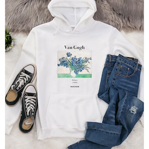 Puede incluir: Sudadera con capucha blanca con una impresión de la pintura "Lirios" de Van Gogh. La sudadera tiene un bolsillo delantero y se combina con vaqueros azules y zapatillas de lona negras. El texto "Van Gogh" e "Irises (1890) TASCHEN" están impresos.